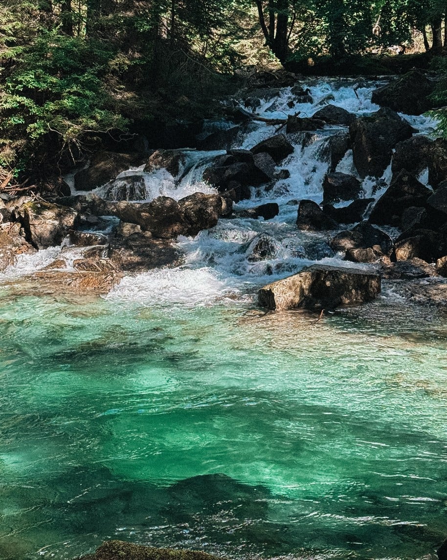 Acqua turchese in pozza davanti a una cascatella che scorre su massi circondati da alberi