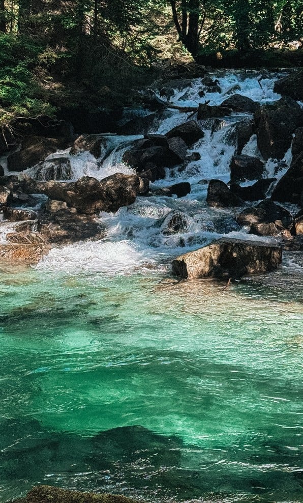 Cascata su massi rocciosi che sfocia in acqua turchese, alberi e ombra sullo sfondo