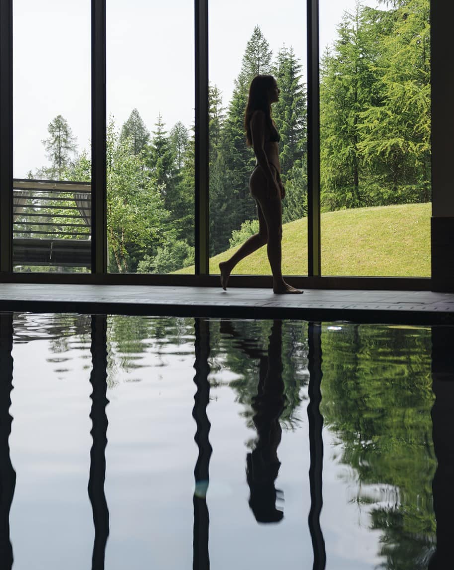 Donna in costume che cammina a bordo piscina interna, vetrate su prato e alberi con riflesso in acqua