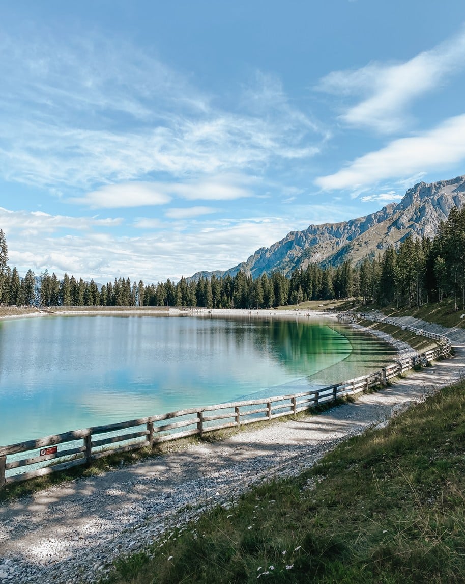 Laghetto alpino turchese con staccionata e sentiero lungo la riva, bosco di conifere e montagne rocciose sullo sfondo