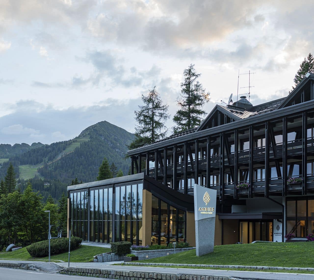 Hotel Ambiez, dall'aspetto moderno con vetrate e balconi, dietro un prato; montagne boschive sullo sfondo
