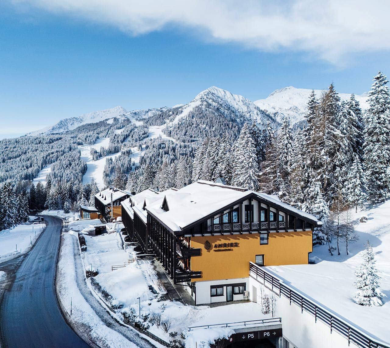 Hotel Ambiez, dall'aspetto moderno con vetrate e balconi, dietro un prato; montagne boschive sullo sfondo