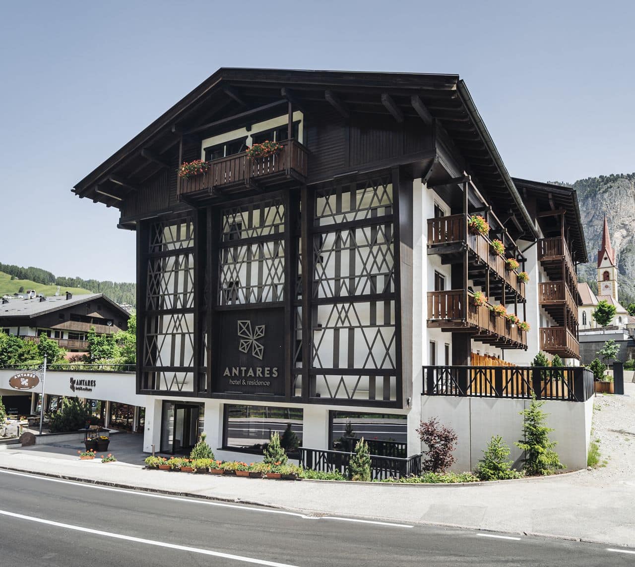 Edificio Antares hotel & residence in stile chalet, balconi con fiori, strada davanti e chiesa sullo sfondo