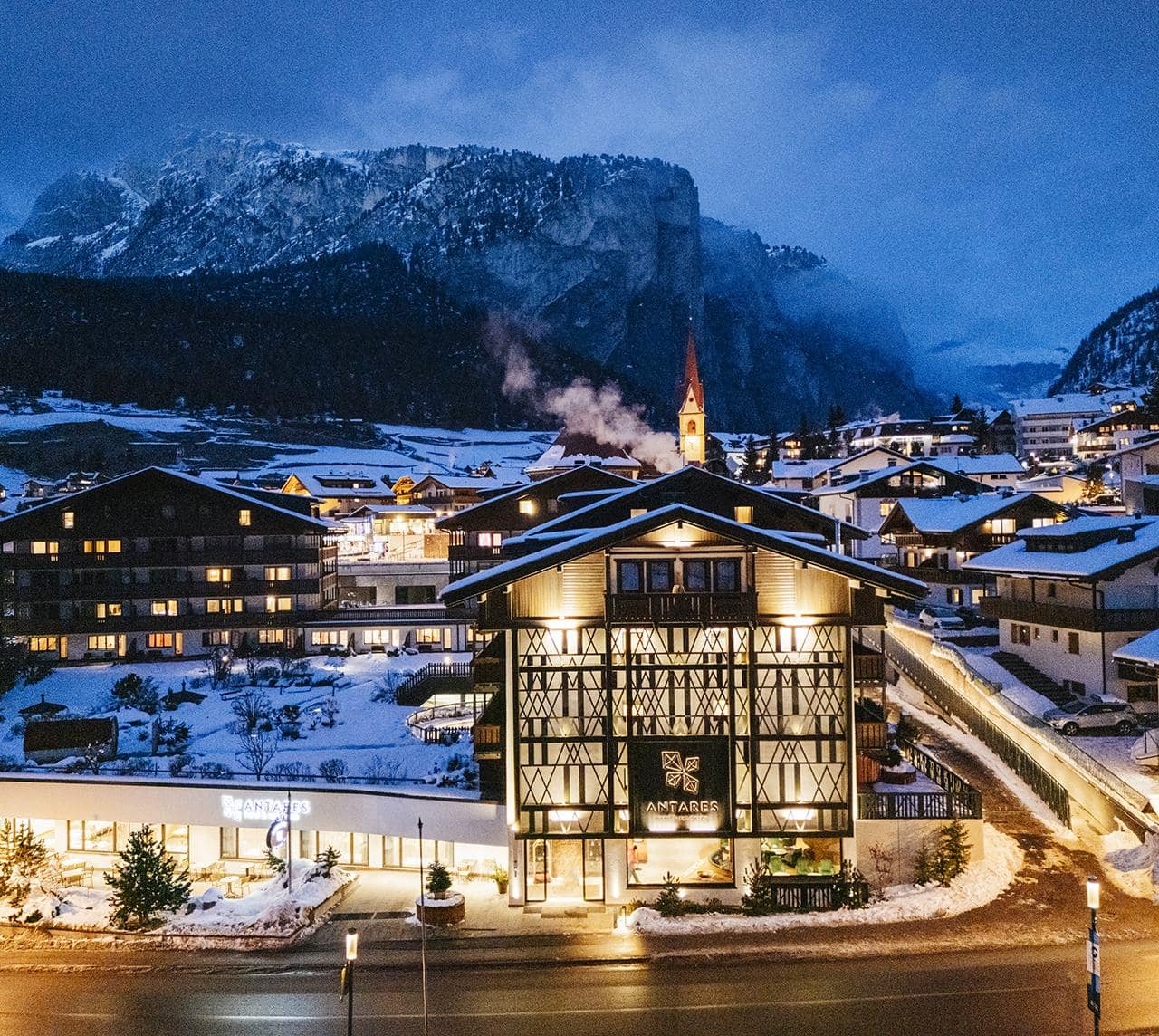 Edificio Antares hotel & residence in stile chalet, balconi con fiori, strada davanti e chiesa sullo sfondo