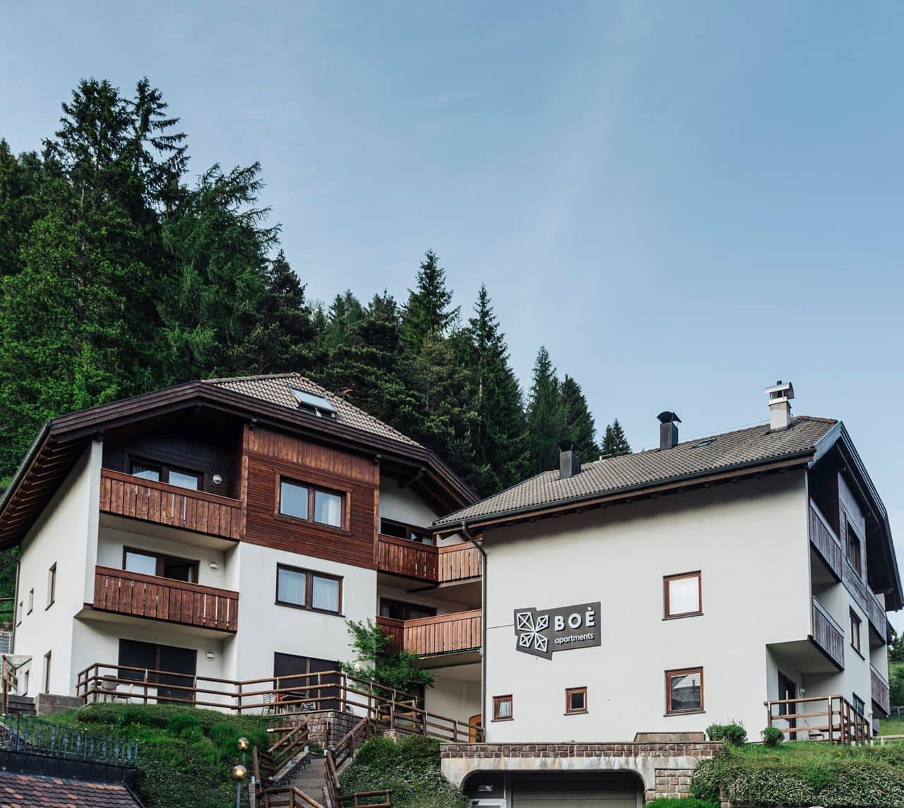 Edificio di BOÈ apartments con balconi in legno, scale esterne e bosco di conifere sullo sfondo