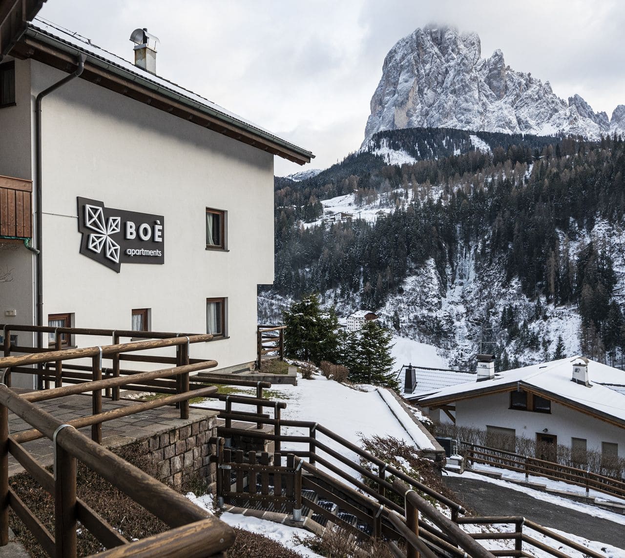 Edificio di BOÈ apartments con balconi in legno, scale esterne e bosco di conifere sullo sfondo