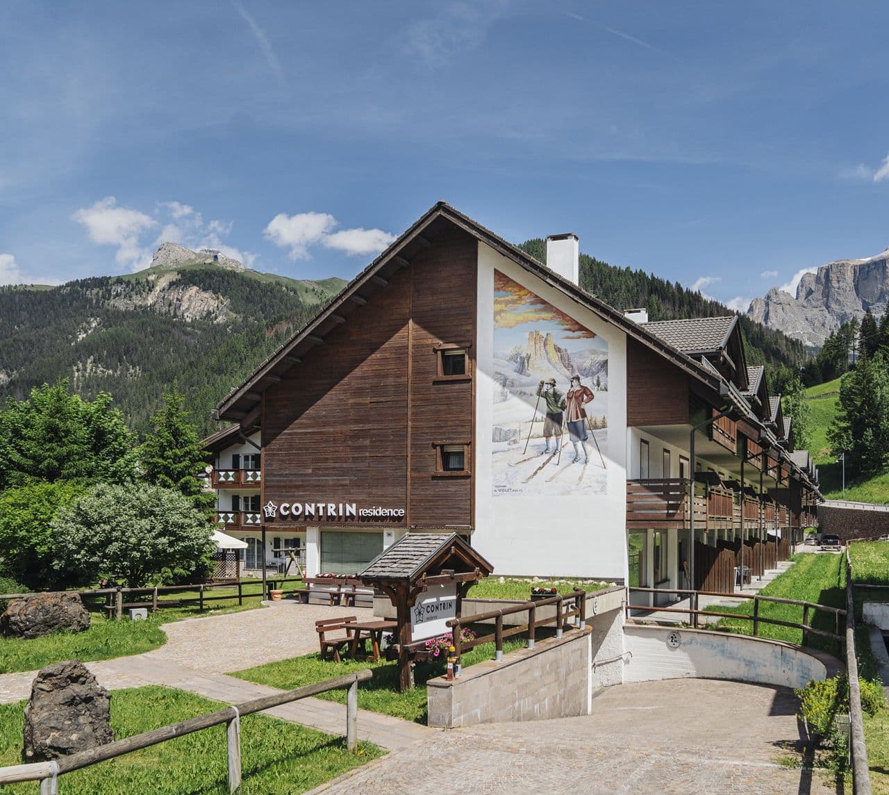 Edificio Contrin residence con murale raffigurante sciatori e montagne, prato e strada ciottolata