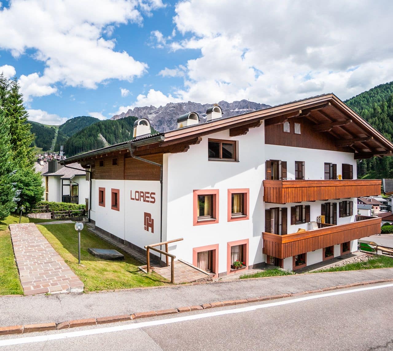 Edificio bianco con balconi in legno e scritta LORES, stradina davanti, montagne e bosco sullo sfondo