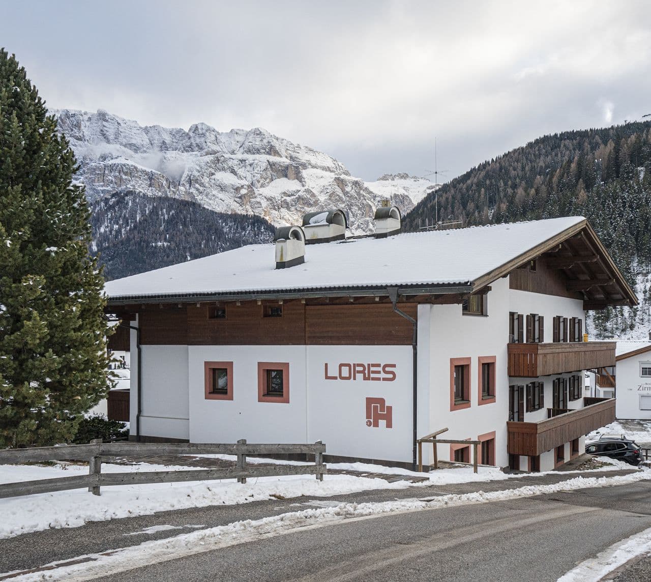 Edificio bianco con balconi in legno e scritta LORES, stradina davanti, montagne e bosco sullo sfondo
