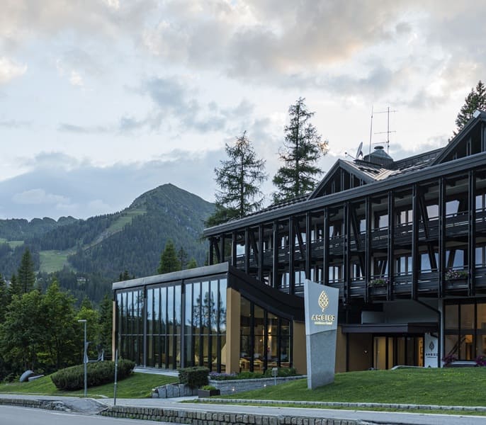 Edificio di Ambiez Residencehotel con vetrate e balconi davanti a pini e montagna sullo sfondo