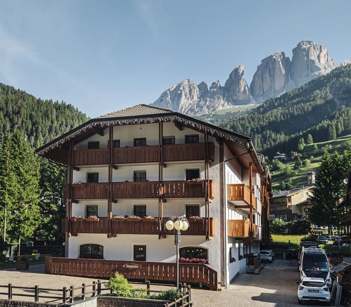 Edificio alberghiero a tre piani con balconi in legno, auto in parcheggio e montagne rocciose sullo sfondo