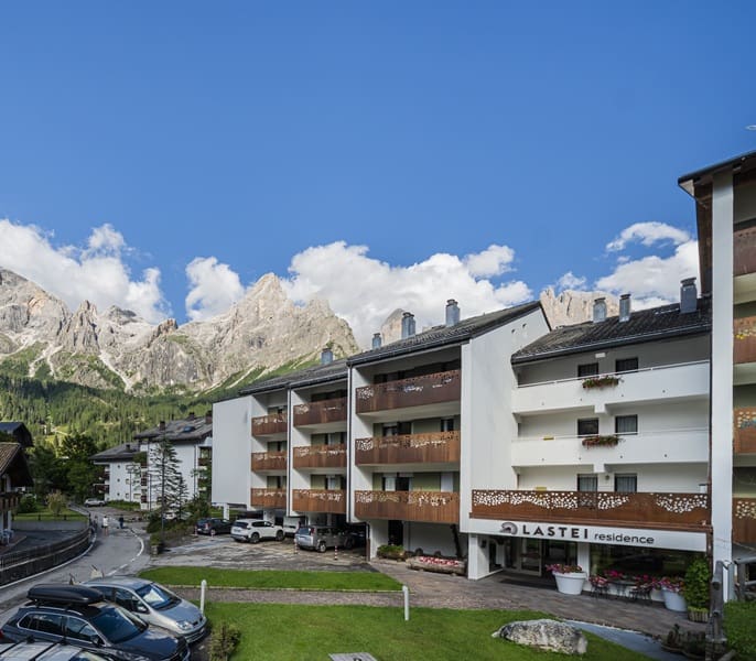 Edificio del residence Lastei con balconi e auto nel parcheggio, montagne rocciose sullo sfondo