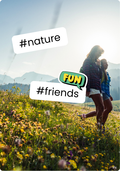 Two women walking in a sunlit wildflower meadow with mountains behind; stickers: #nature #friends FUN