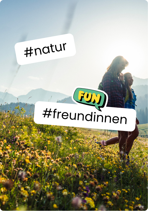 Zwei Freundinnen spazieren lachend durch eine blühende Bergwiese vor sonnigen Bergen