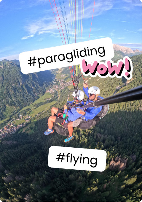 Two people tandem paragliding over forested mountains and a valley, selfie pole visible