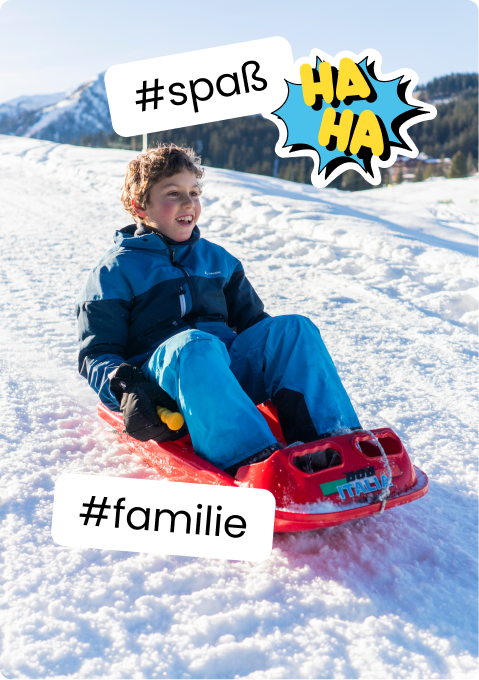 Junge in blauer Winterjacke lacht auf rotem Schlitten im Schnee; Texte #spaß und #familie.