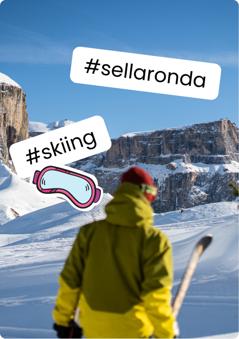 Person in yellow jacket and red hat holding skis, facing snow-covered mountains; stickers reading #sellaronda #skiing