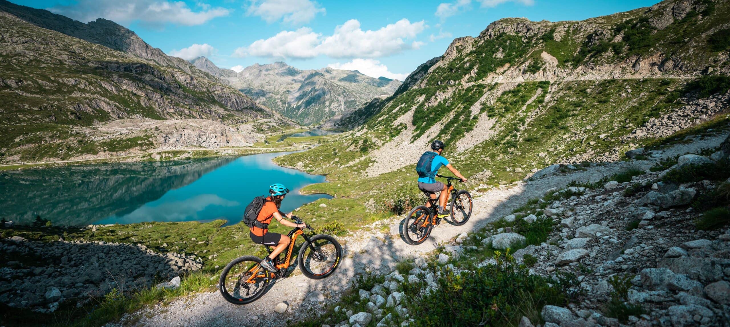 Due ciclisti in mountain bike su un sentiero roccioso che costeggia un lago alpino circondato da montagne.