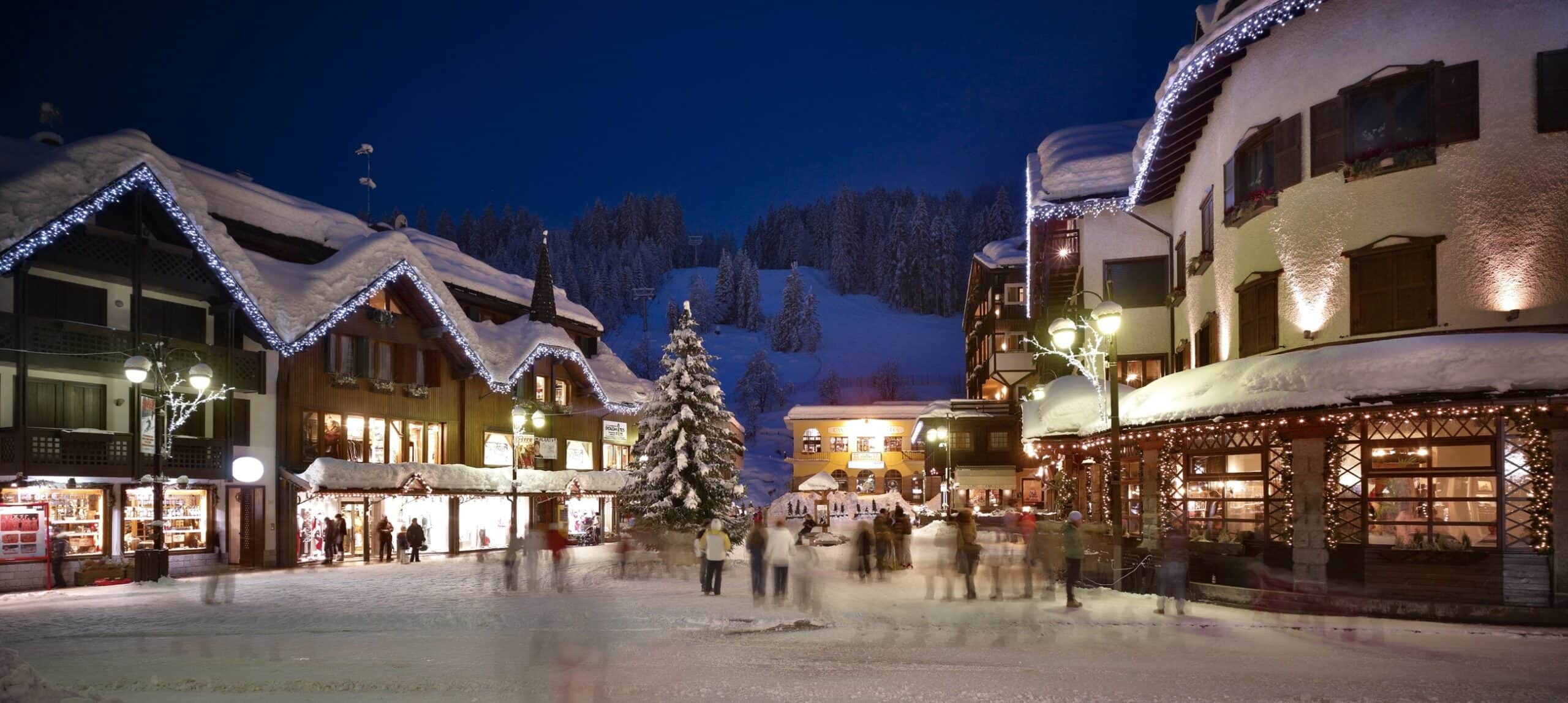 Piazza innevata serale con grande abete illuminato, edifici decorati e persone in movimento