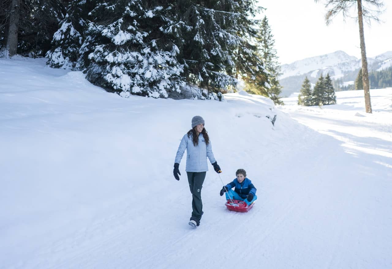 Donna che tira un bambino su una slitta rossa lungo un sentiero innevato, alberi e montagne sullo sfondo