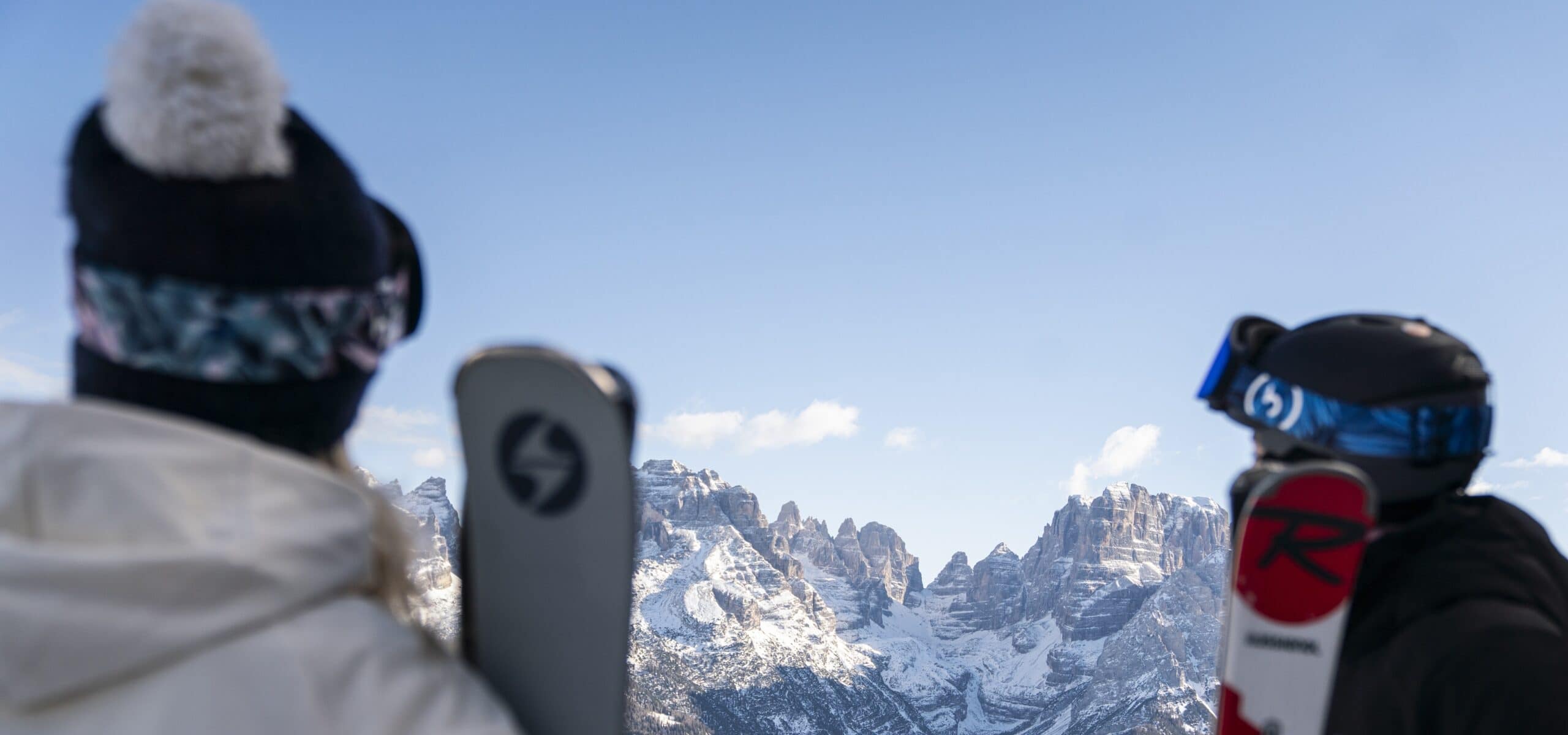 Due persone in abbigliamento da sci, viste di spalle, guardano montagne innevate sotto cielo azzurro