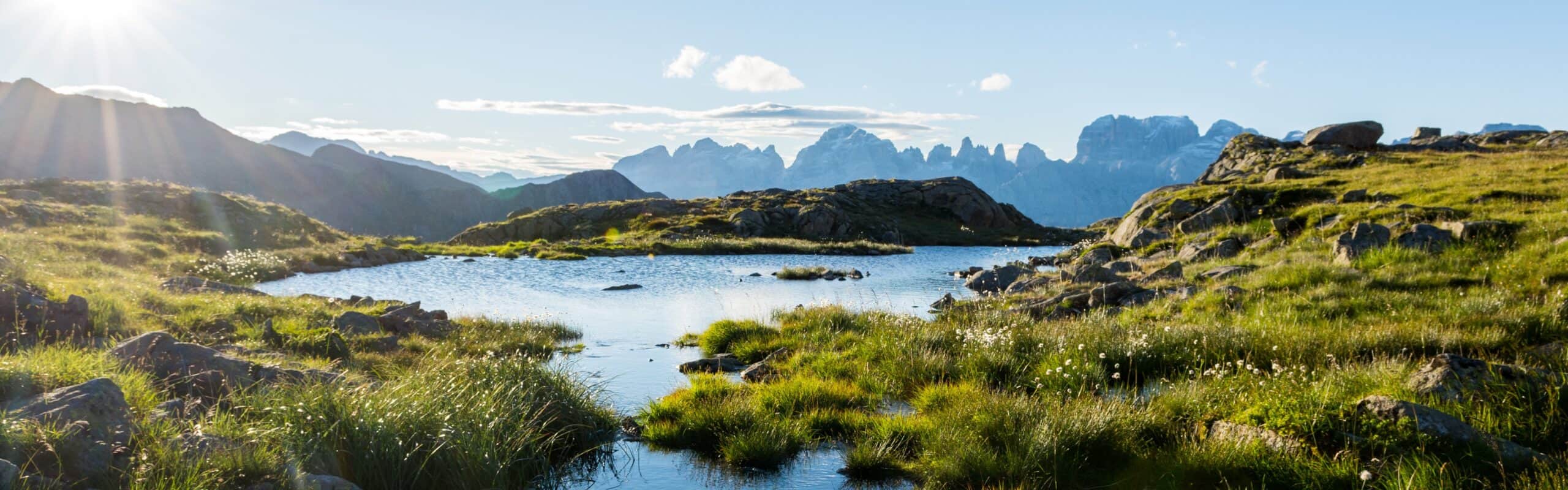 Laghetto alpino tra prati e massi, acqua calma con riflessi, catena di montagne sullo sfondo e cielo sereno.