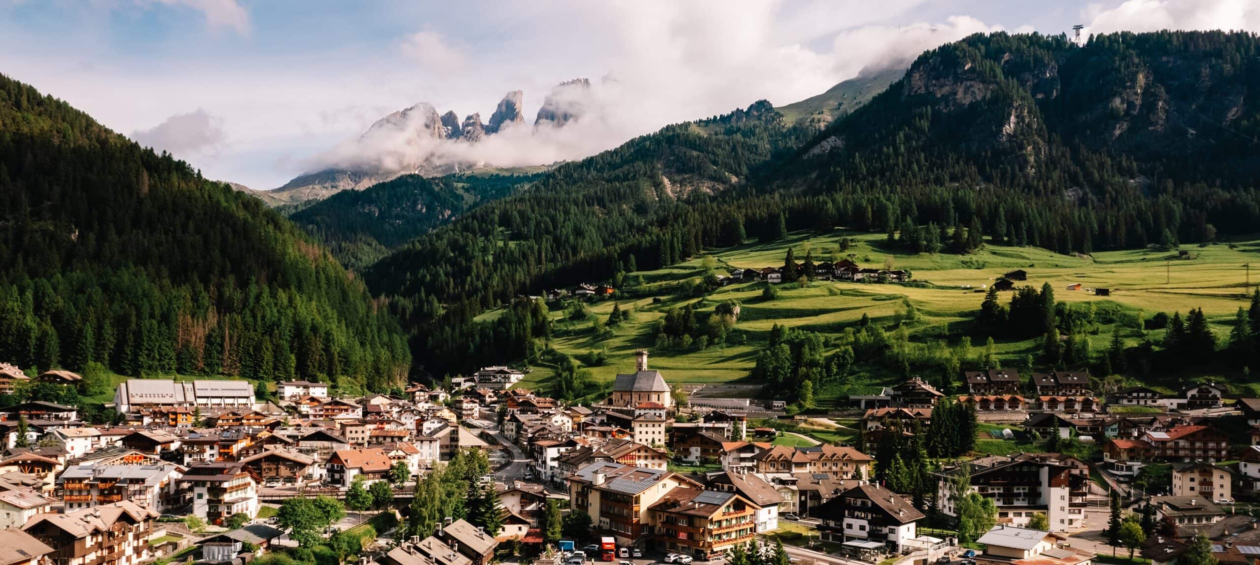 Paese alpino con chiesa e case ai piedi di colline verdi e monti boscosi avvolti dalla nebbia