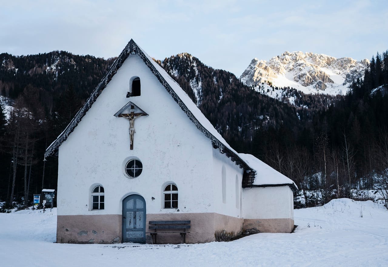 Piccola chiesa bianca con crocifisso sulla facciata, panca davanti e paesaggio montano innevato dietro