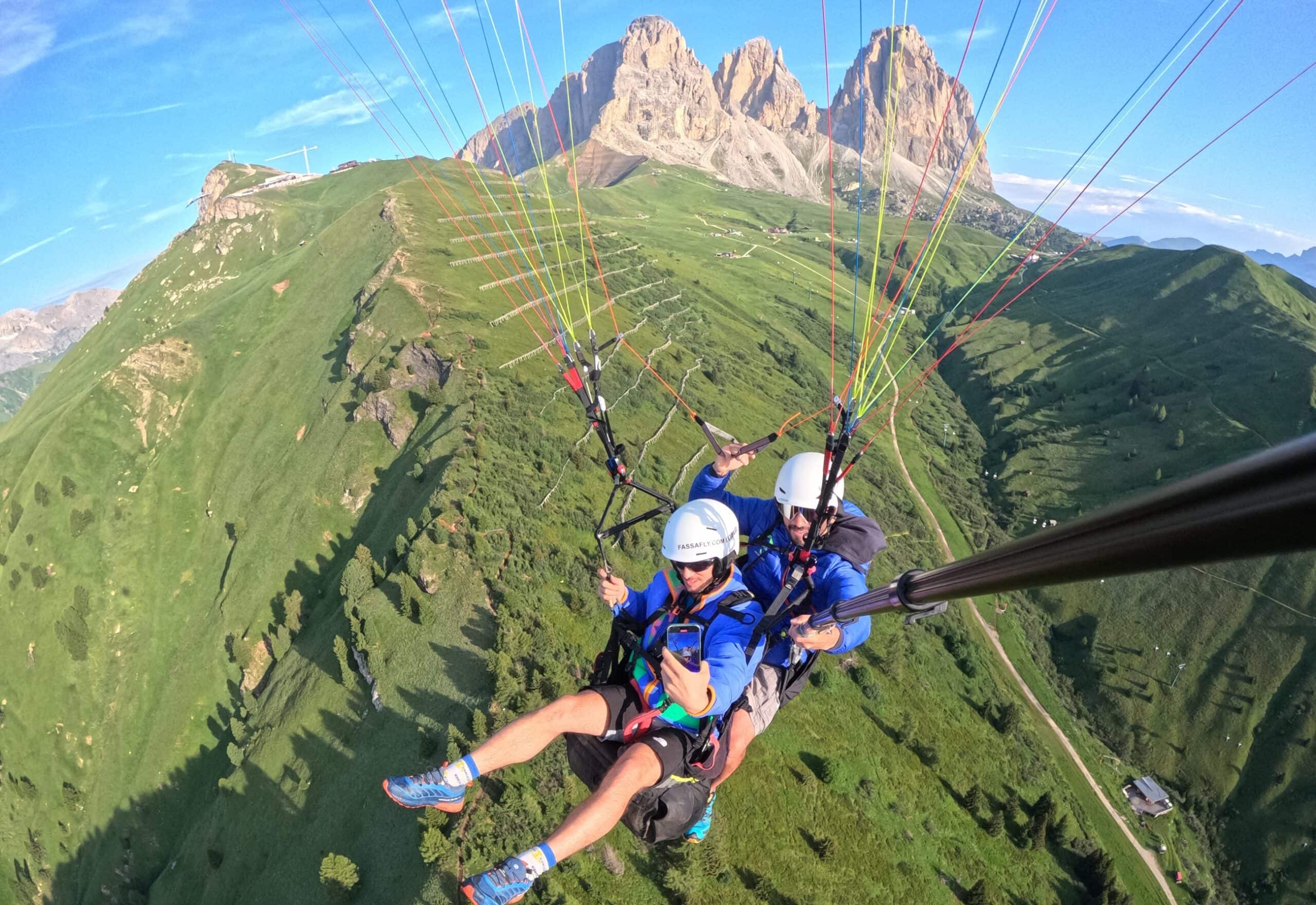 Due persone in tandem parapendio sospese sopra pascoli verdi, selfie con bastone e montagne rocciose sullo sfondo