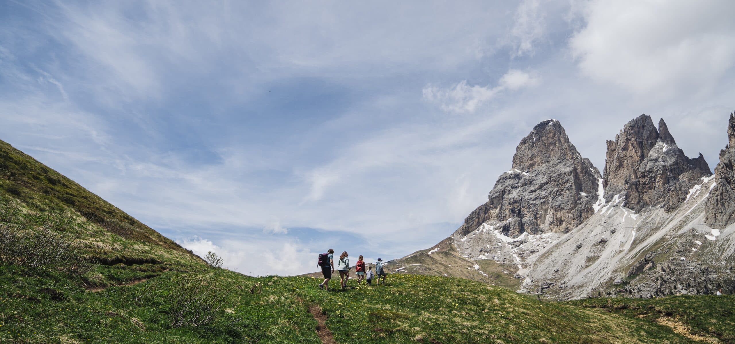 Gruppo di escursionisti cammina su prato di montagna verso ripide pareti rocciose sotto cielo nuvoloso