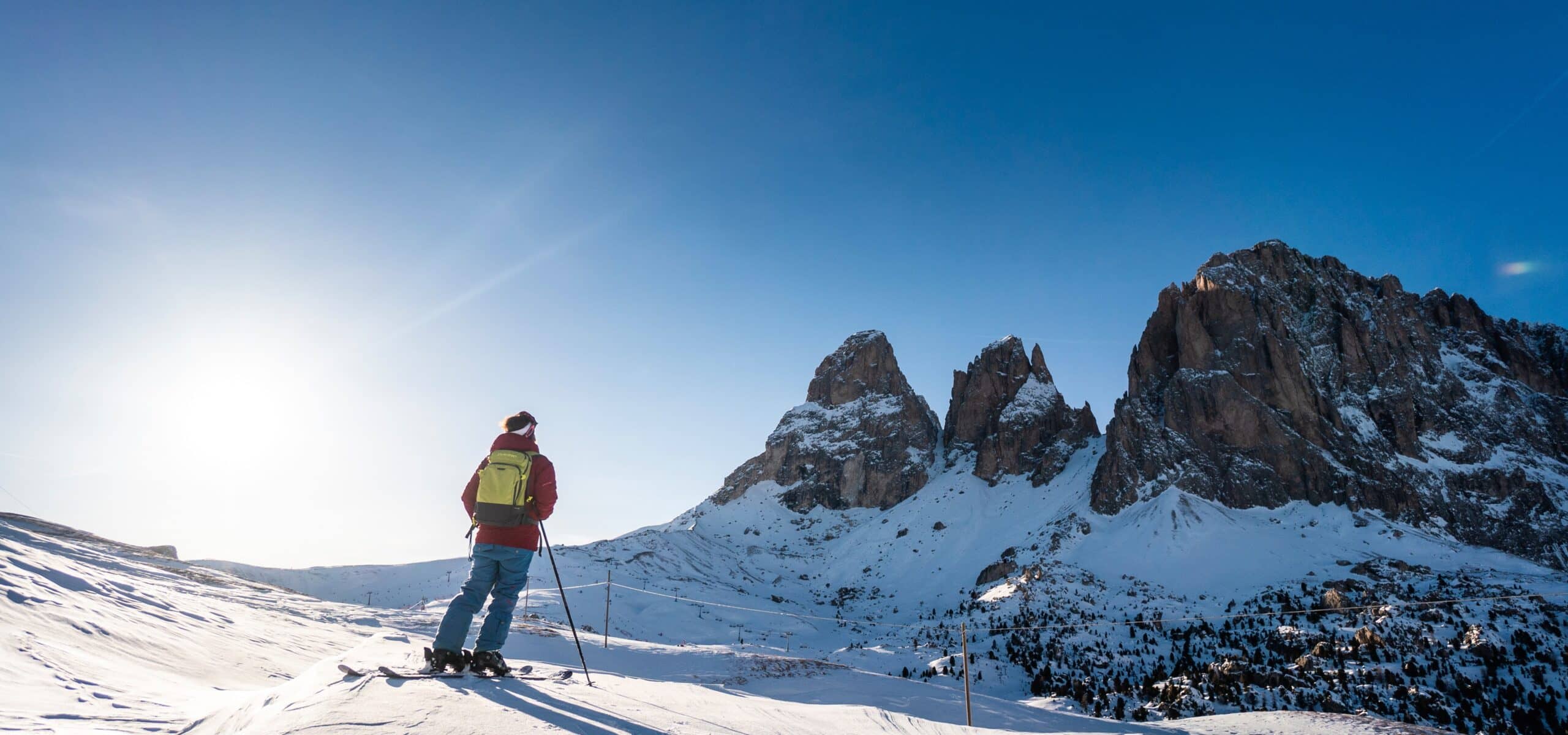 Sciatore con zaino in piedi su pendio innevato guarda alte pareti rocciose innevate sotto cielo azzurro