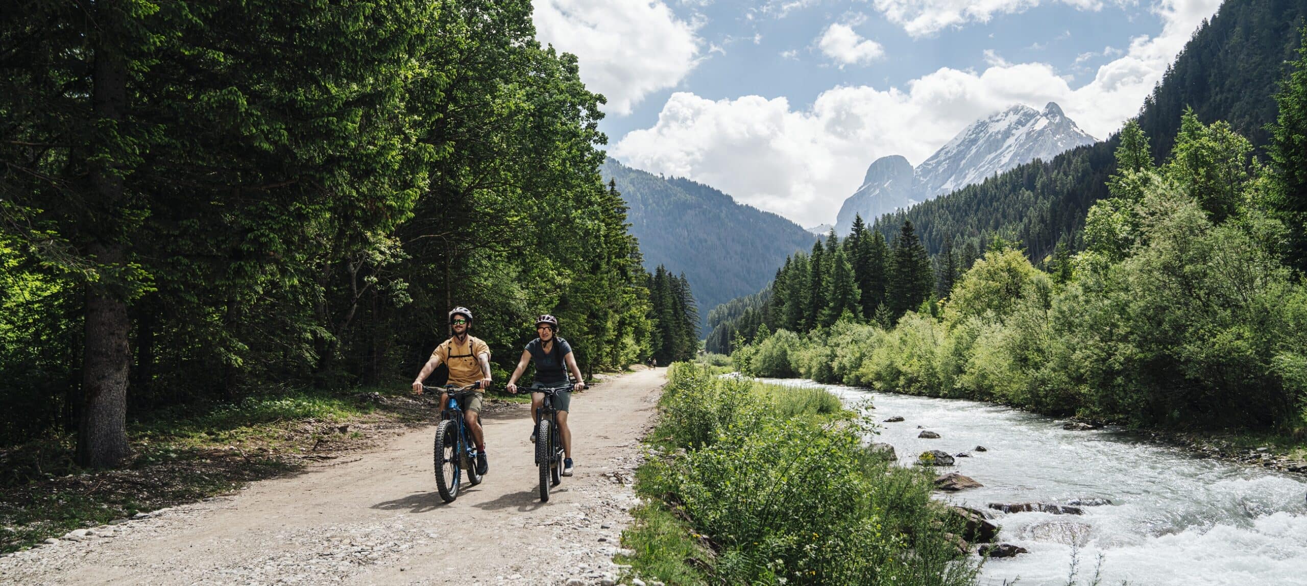 Due ciclisti in mountain bike su sentiero accanto a un fiume, foresta e montagne innevate sullo sfondo