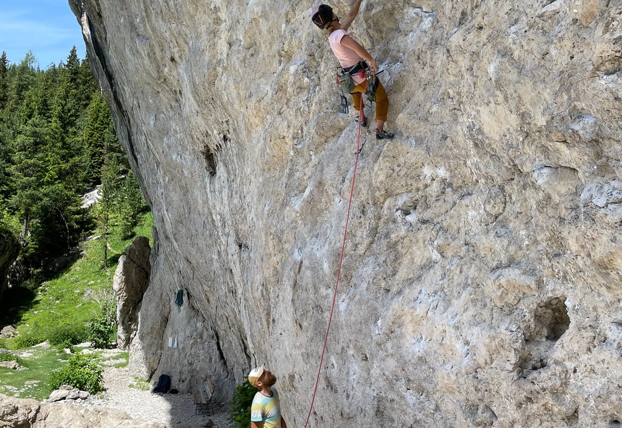 Arrampicatore su parete rocciosa assicurato con corda rossa, persona a terra lo assicura; bosco sullo sfondo.