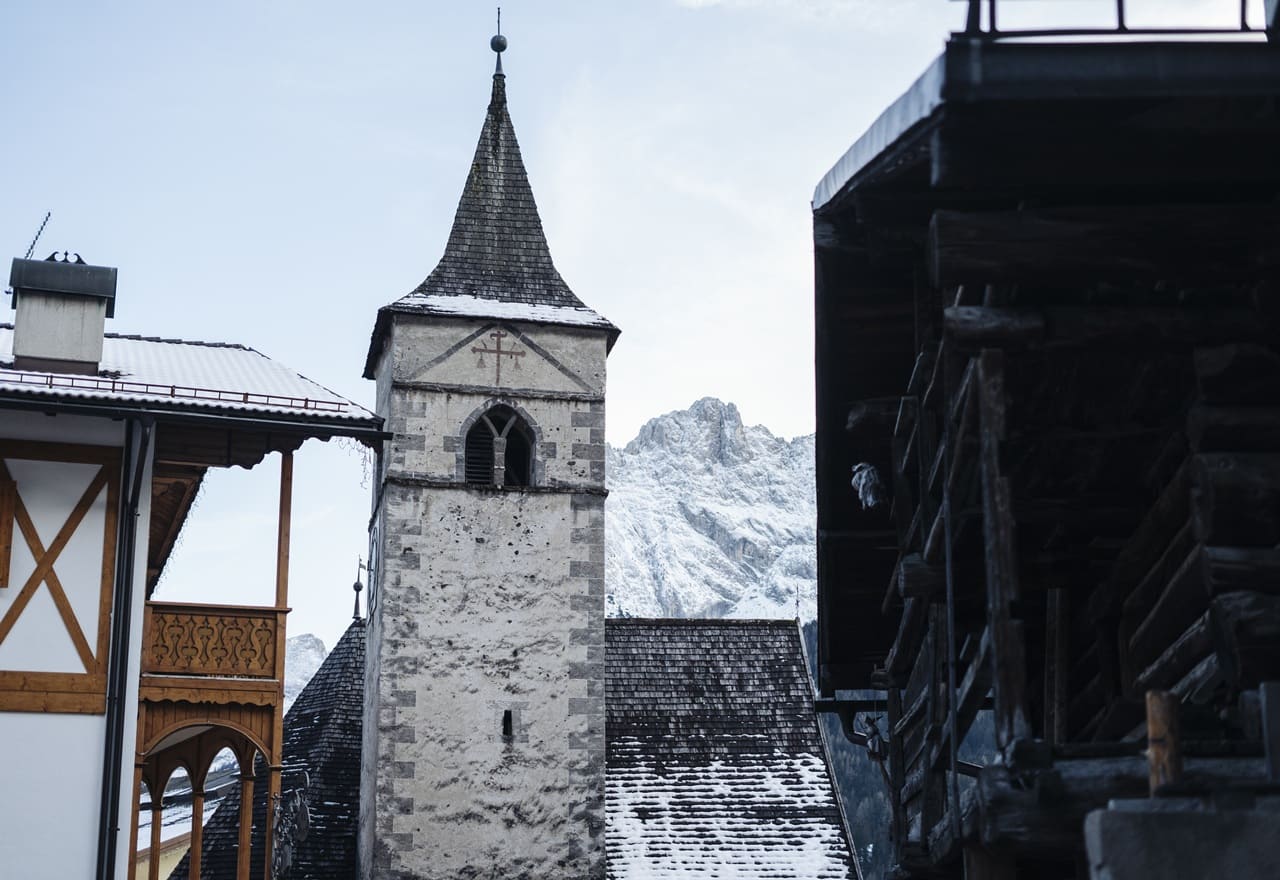 Campanile di chiesa in pietra con tetto a punta, case in legno ai lati e montagna innevata sullo sfondo