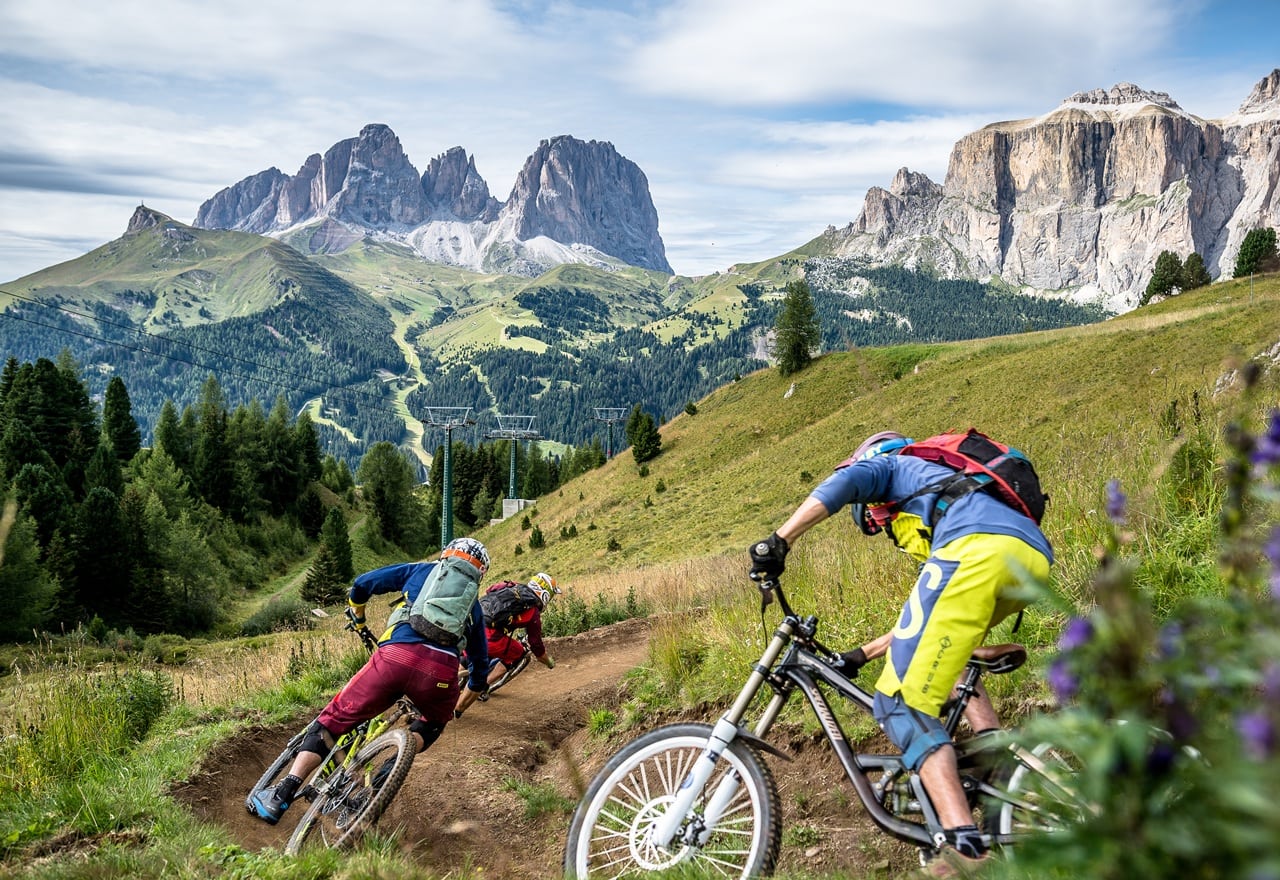 Tre ciclisti in discesa su sentiero alpino tra prati e boschi, con alte cime rocciose sullo sfondo