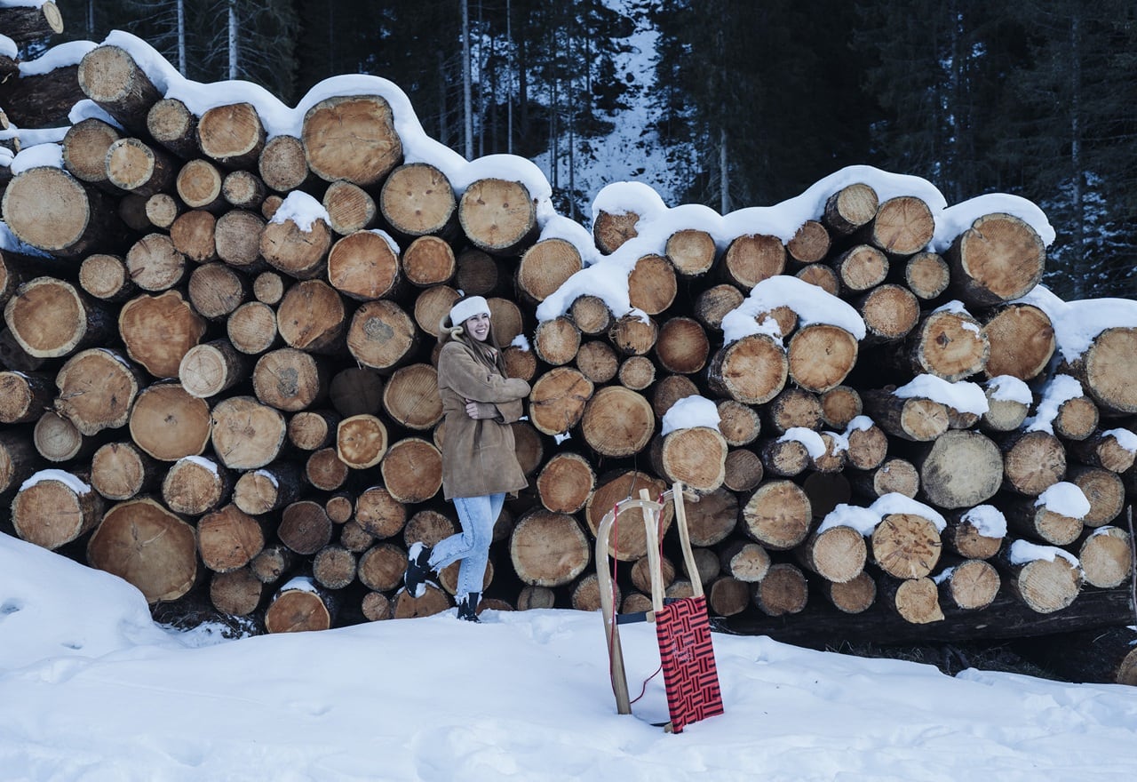 Donna con cappotto e berretto appoggiata a una pila di tronchi innevati, slitta in piedi sulla neve