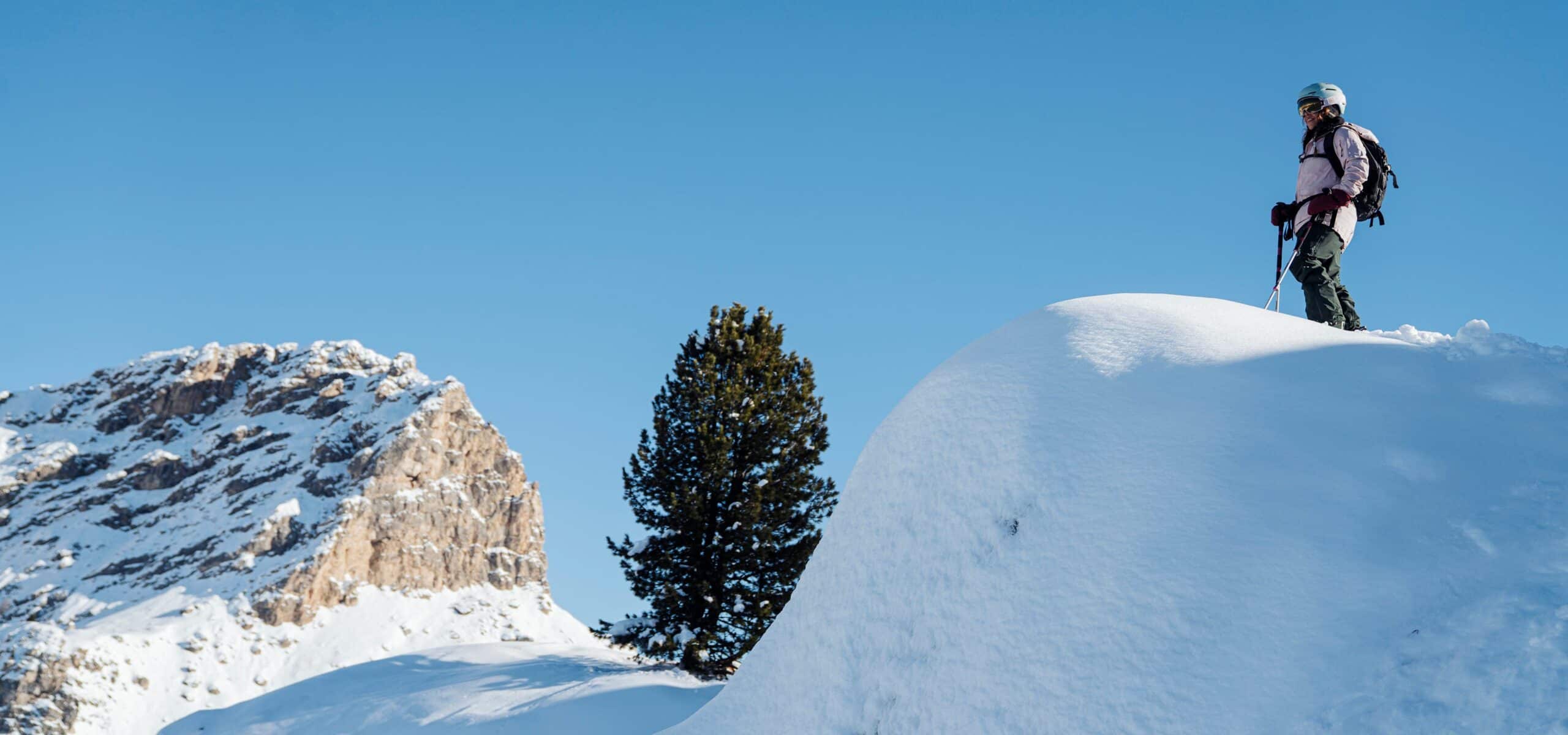 Sciatore in piedi su un dosso innevato con bastoncini, albero e parete rocciosa sullo sfondo