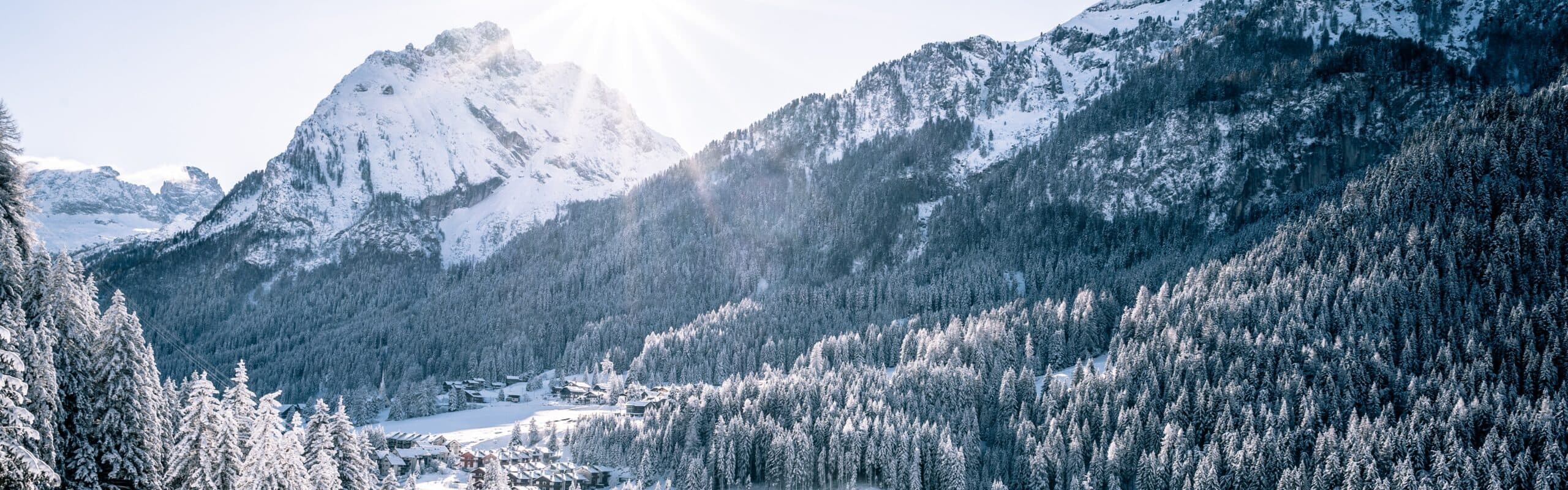 Paese innevato in valle, circondato da boschi di conifere e montagne ricoperte di neve, raggio di sole sul picco