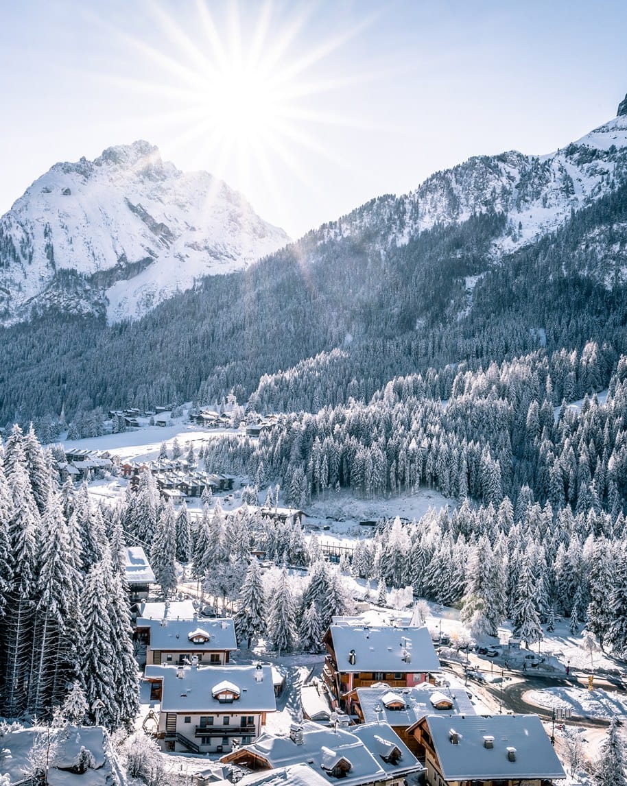 Paese alpino innevato con chalet, foresta di conifere e montagne innevate illuminate dal sole
