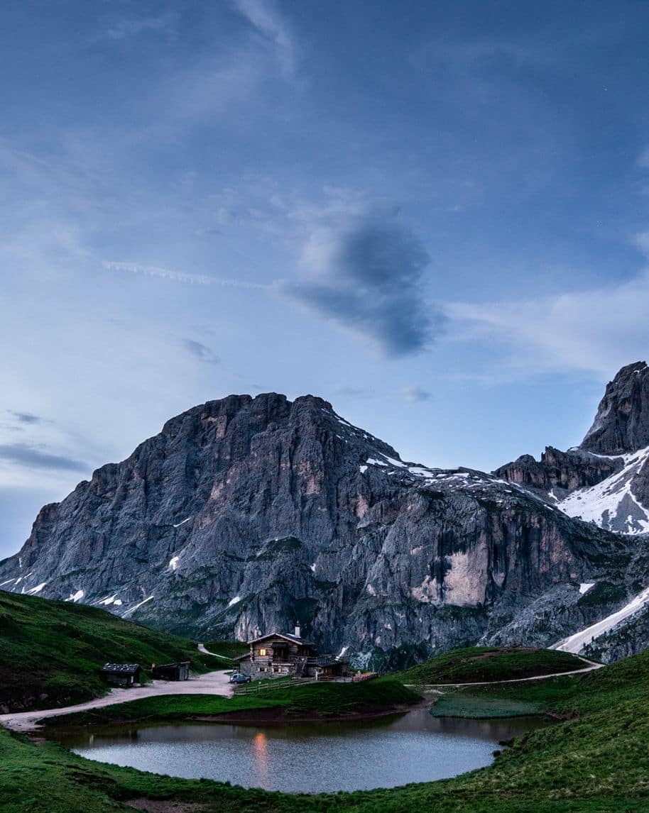 Baita illuminata accanto a un laghetto alpino, davanti a una parete rocciosa al crepuscolo