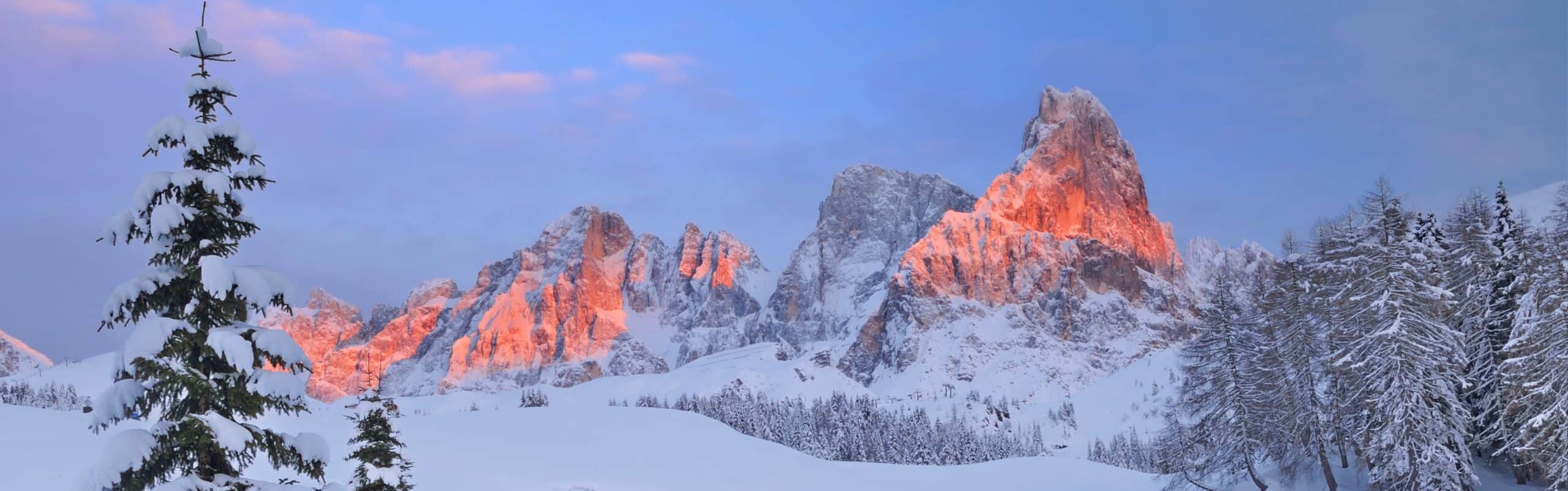 Catena montuosa innevata illuminata dal sole arancione, alberi con neve in primo piano