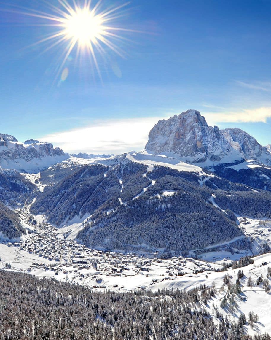 Paese innevato nella valle con boschi e pendii, massiccio roccioso sullo sfondo e sole brillante