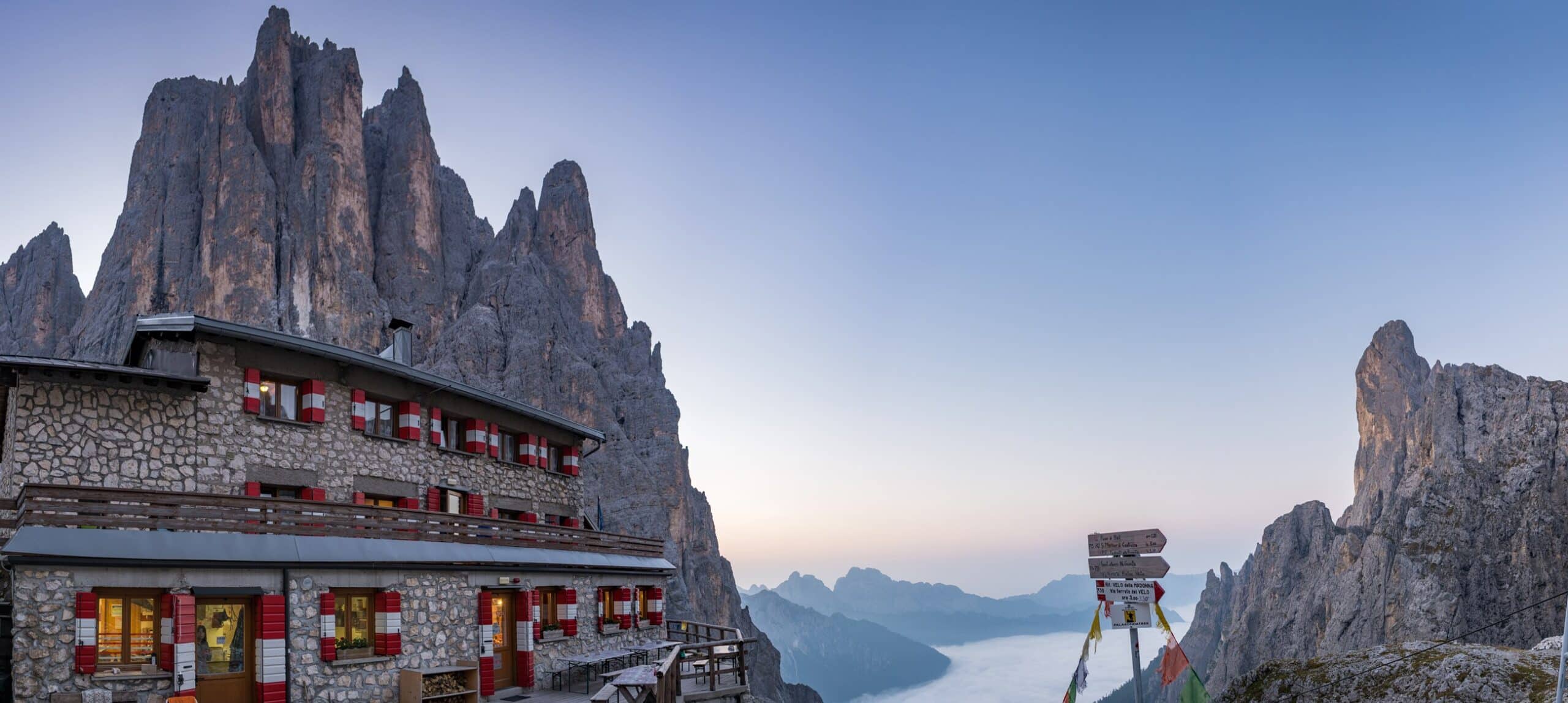 Rifugio alpino in pietra con persiane rosse, picchi rocciosi sullo sfondo e valle nebbiosa con cartelli