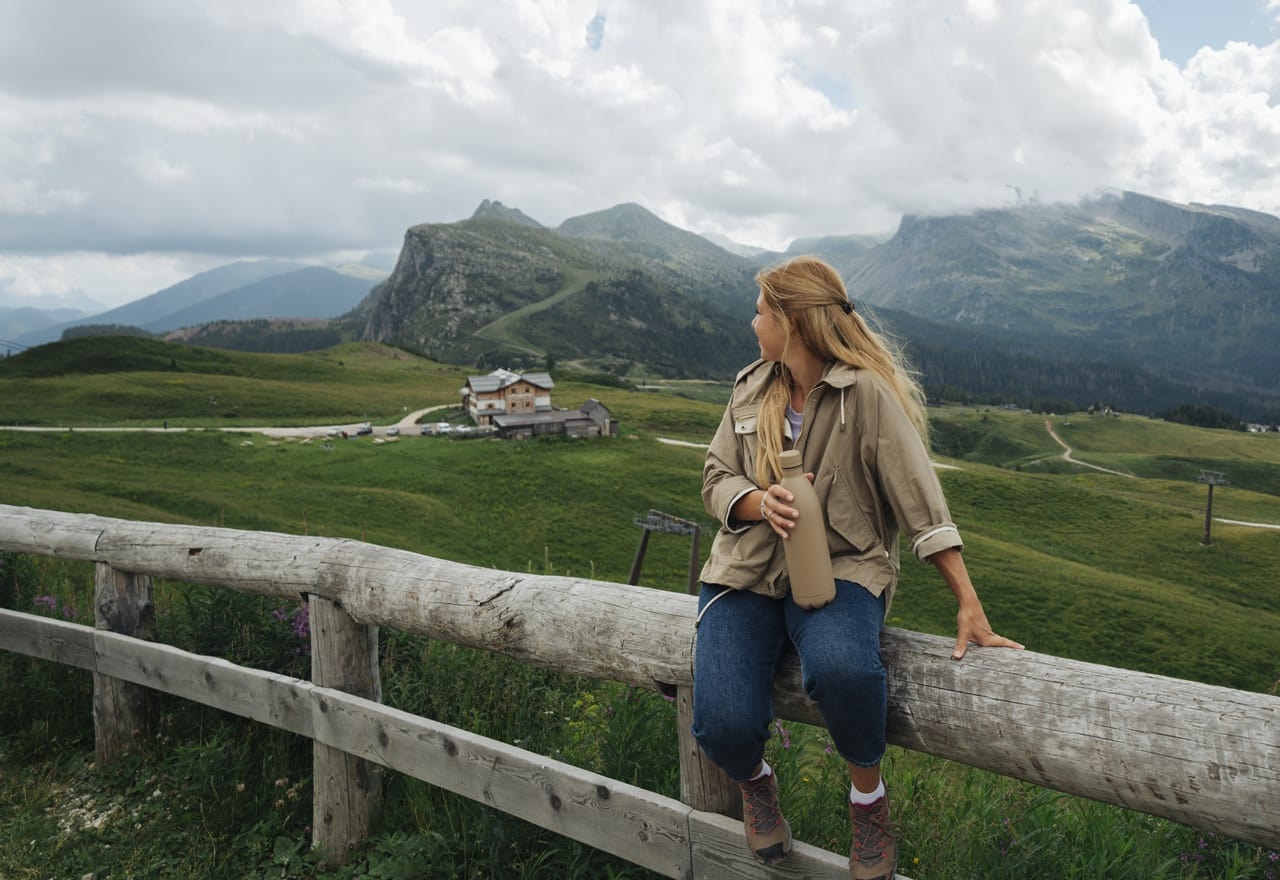Persona seduta su una staccionata in legno con borraccia, guarda vallata verde e montagne nuvolose.