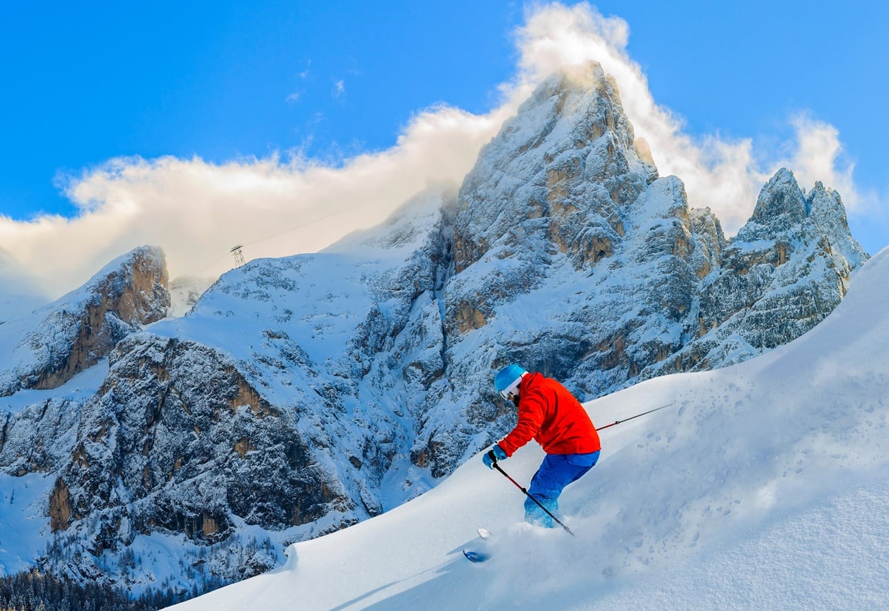 Sciatore con giacca rossa e casco blu scende su neve fresca davanti a alte montagne rocciose innevate