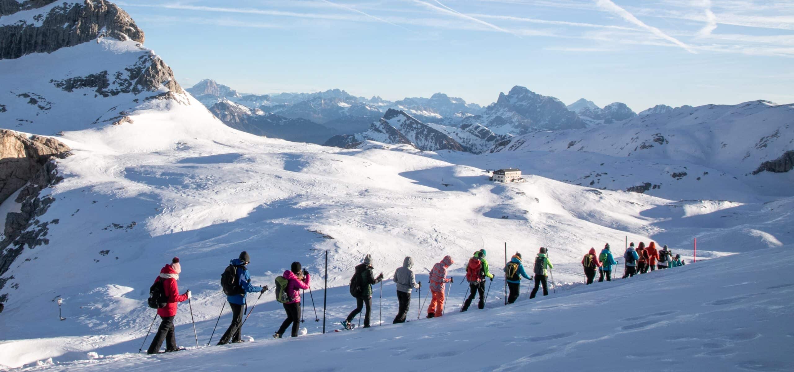 Gruppo di escursionisti con ciaspole e bastoncini in fila su pendio innevato, montagne e rifugio sullo sfondo