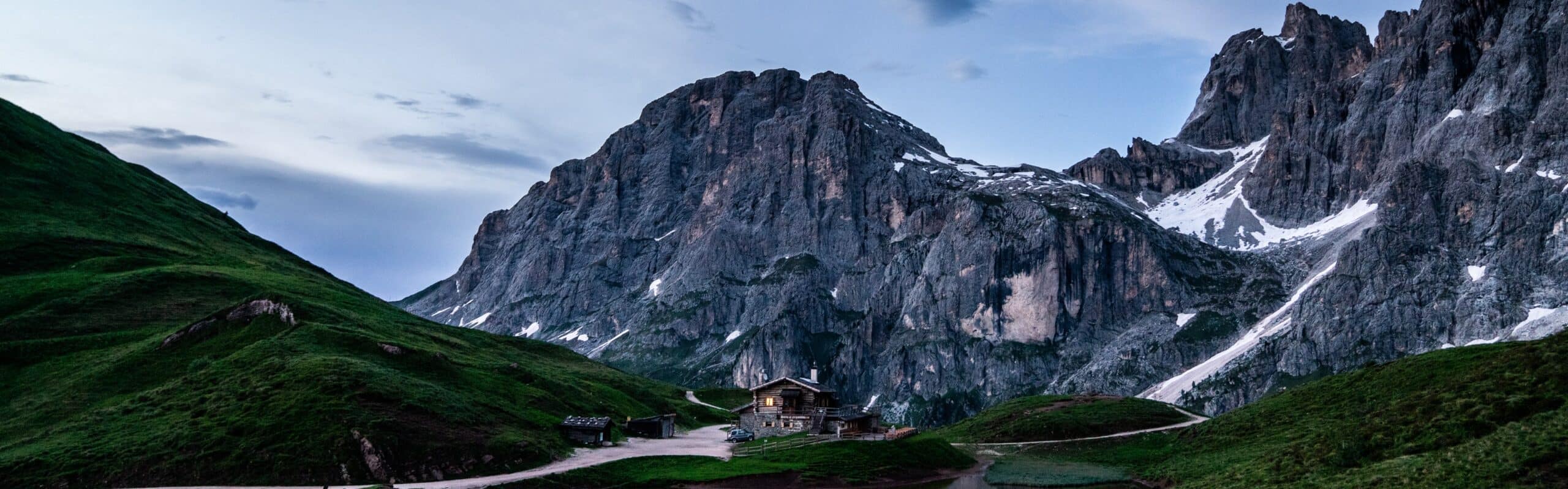 Baita isolata con luce alla finestra in valle erbosa, sentiero e alta parete rocciosa con chiazze di neve.