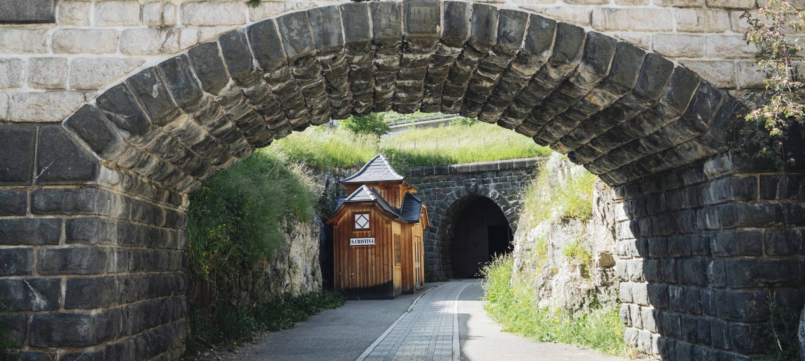 Arco in pietra incornicia sentiero con casetta di legno con targa 'S.CRISTINA' e ingresso di tunnel