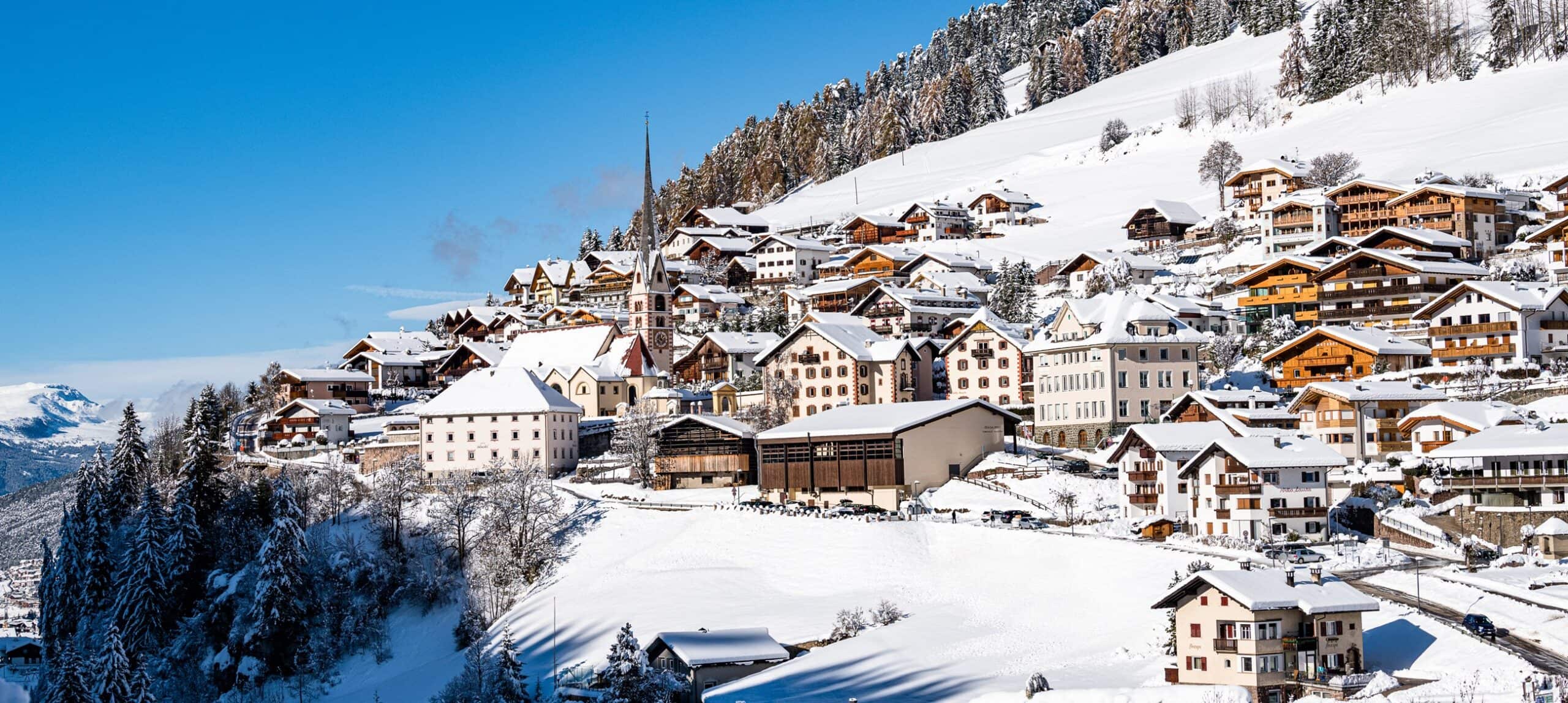 Borgo alpino innevato su pendio con case e alta chiesa a punta, alberi coperti di neve e cielo azzurro