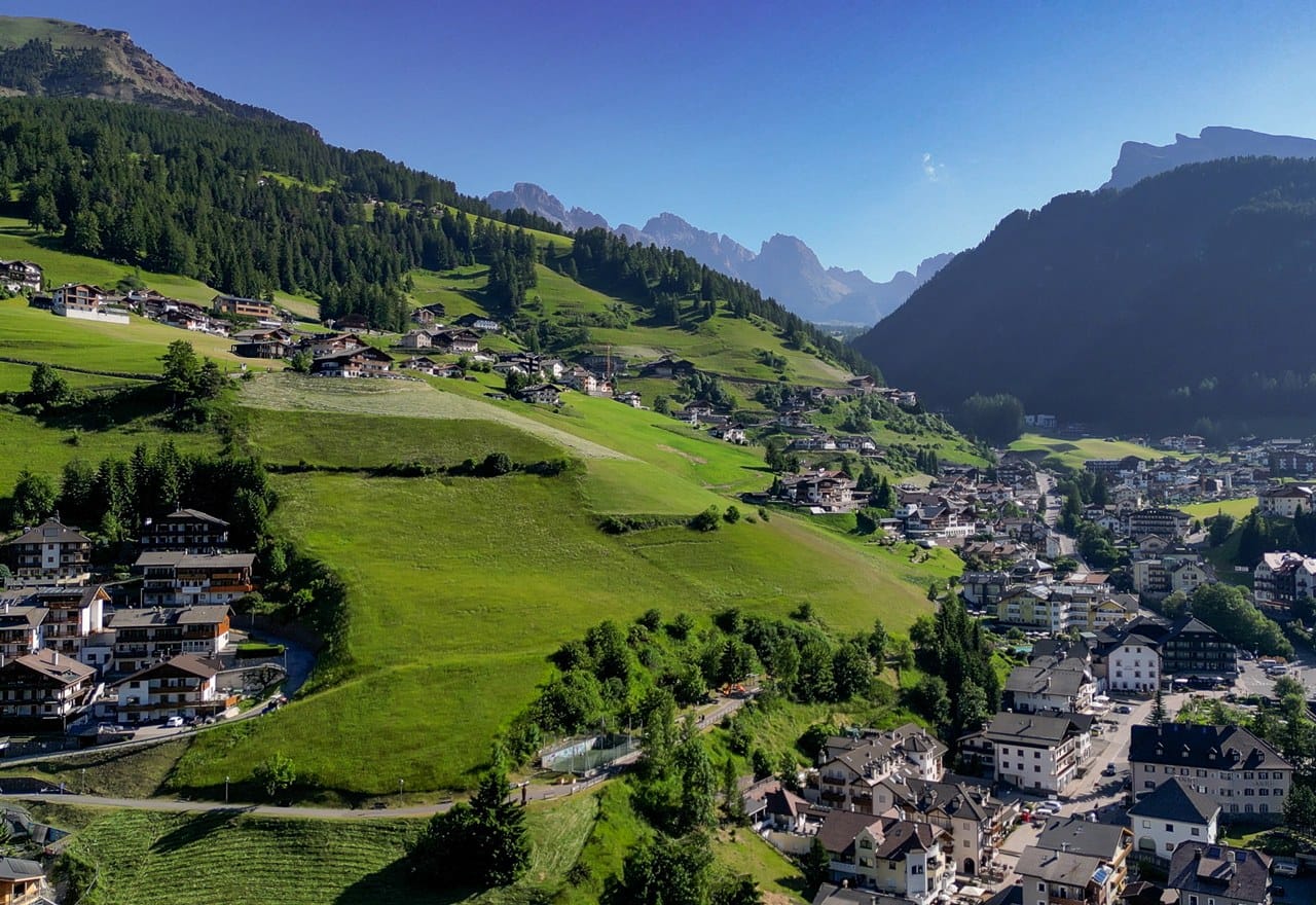 Paese alpino su pendii erbosi con case sparse, foresta e cime montuose sullo sfondo.